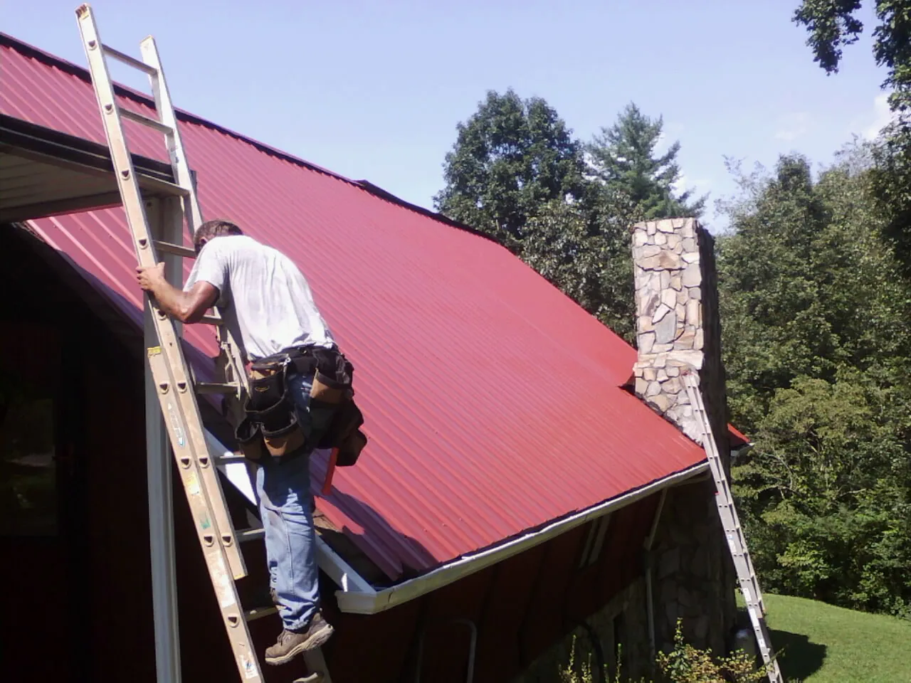 Roofer on a ladder beside a red metal roof and stone chimney — Soffit & Fascia Repair by Roofing Brothers in Carversville